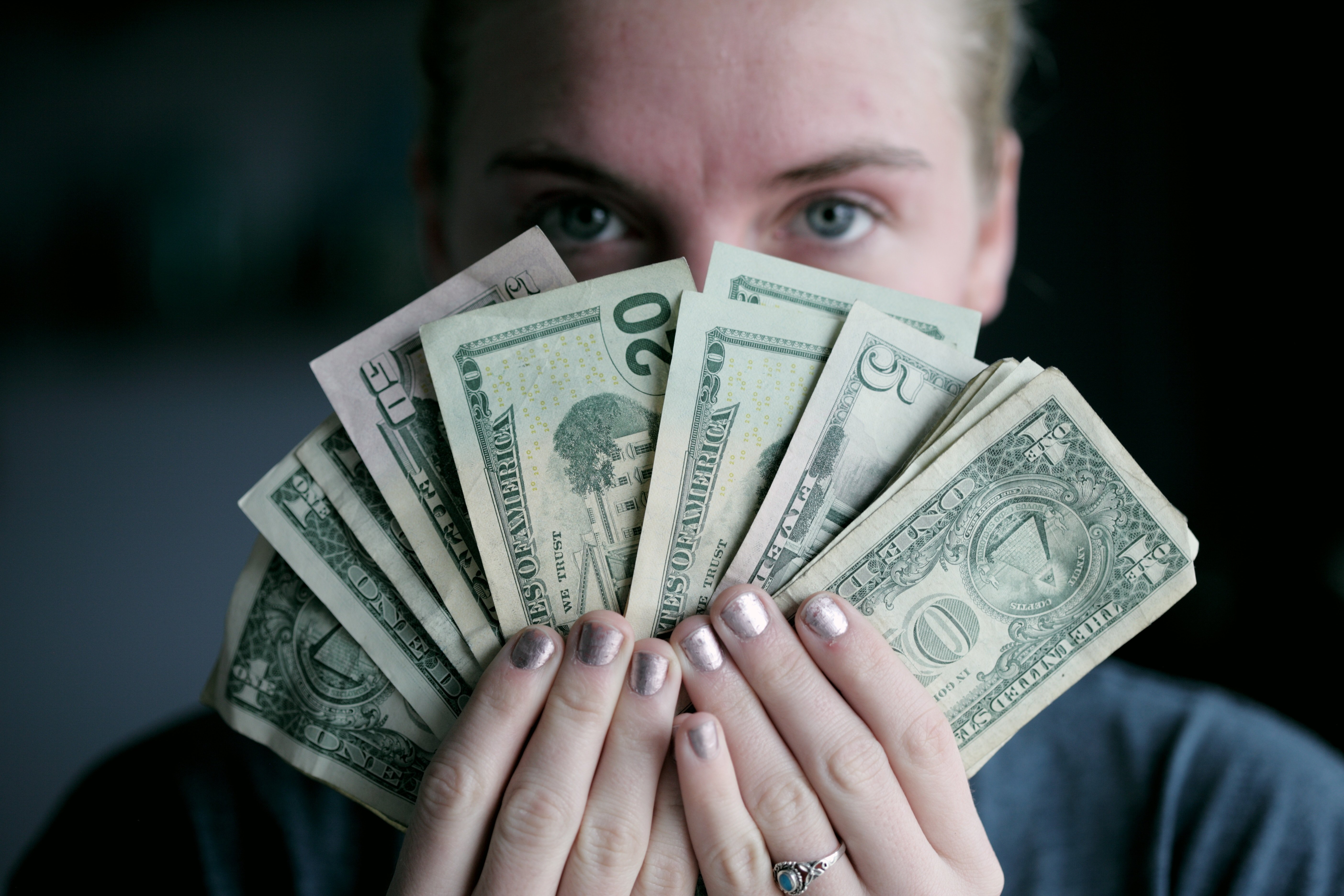 Person holding different denominations of US Currency with two hands covering their face while looking at you